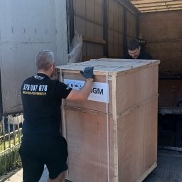 VERTIGO PRZEPROWADZKI Michał Rajczak - Pracownik w czarnej koszulce z logo firmy transportowej przesuwa dużą, drewnianą skrzynię do wnętrza ciężarówki. W tle widoczny drugi pracownik.
