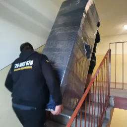 Dwóch mężczyzn w czarnych bluzach wnosi po schodach duży, owinięty mebel. Na bluzie widoczny numer telefonu. Typowe ujęcie z przeprowadzki.