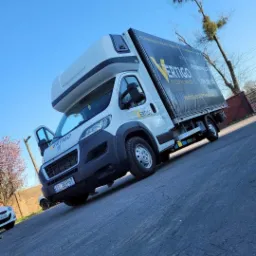 Biały samochód dostawczy z otwartymi drzwiami kierowcy, z logo firmy transportowej 'VERTIGO' na boku plandeki i na karoserii, tablica rejestracyjna EL 3EP67, sfotografowany pod kątem na tle...