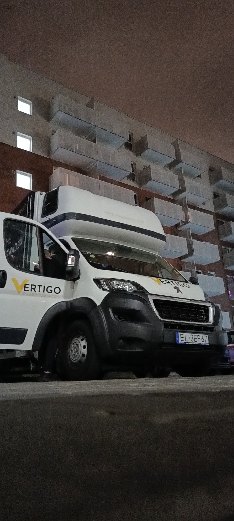 Biała furgonetka z otwartymi drzwiami na tle bloku mieszkalnego, logo firmy przeprowadzkowej Vertigo widoczne na boku pojazdu, nocne oświetlenie.