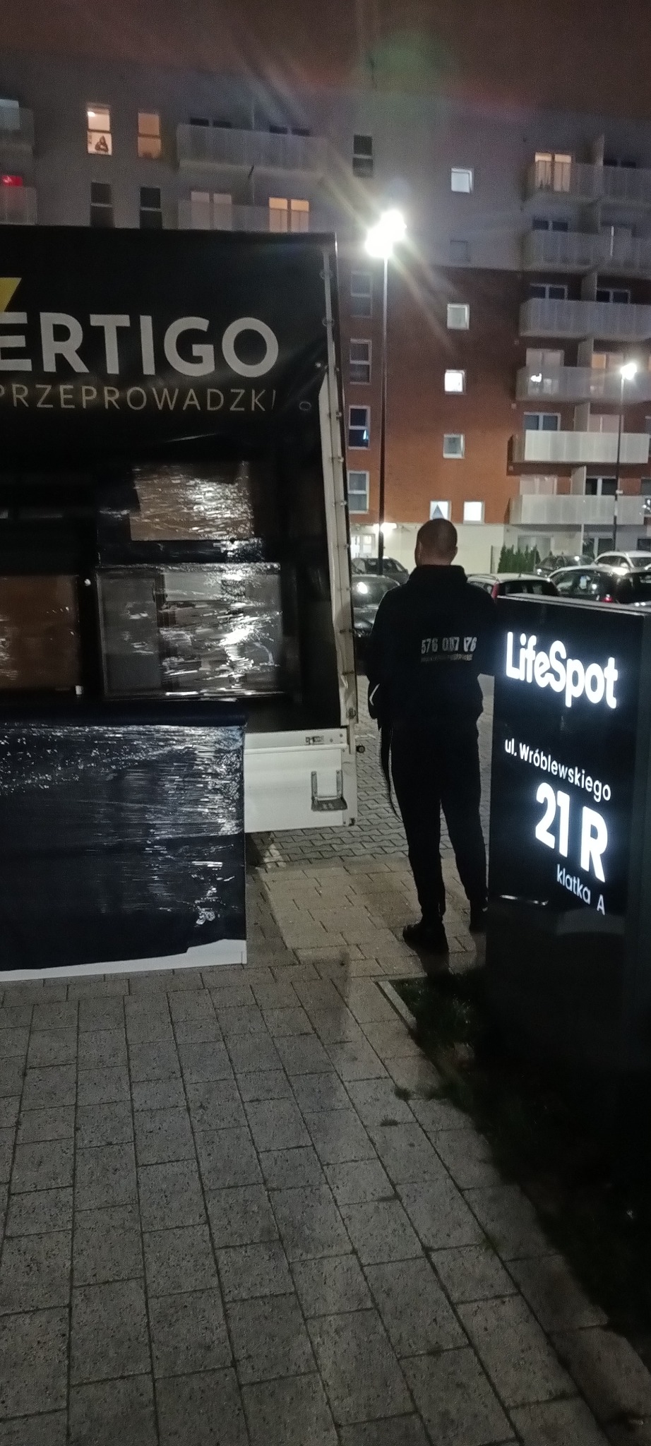 Ciężarówka firmy Vertigo Przeprowadzki z załadunkiem mebli w folii, na tle bloku mieszkalnego. Obok stoi mężczyzna i tablica z adresem LifeSpot.