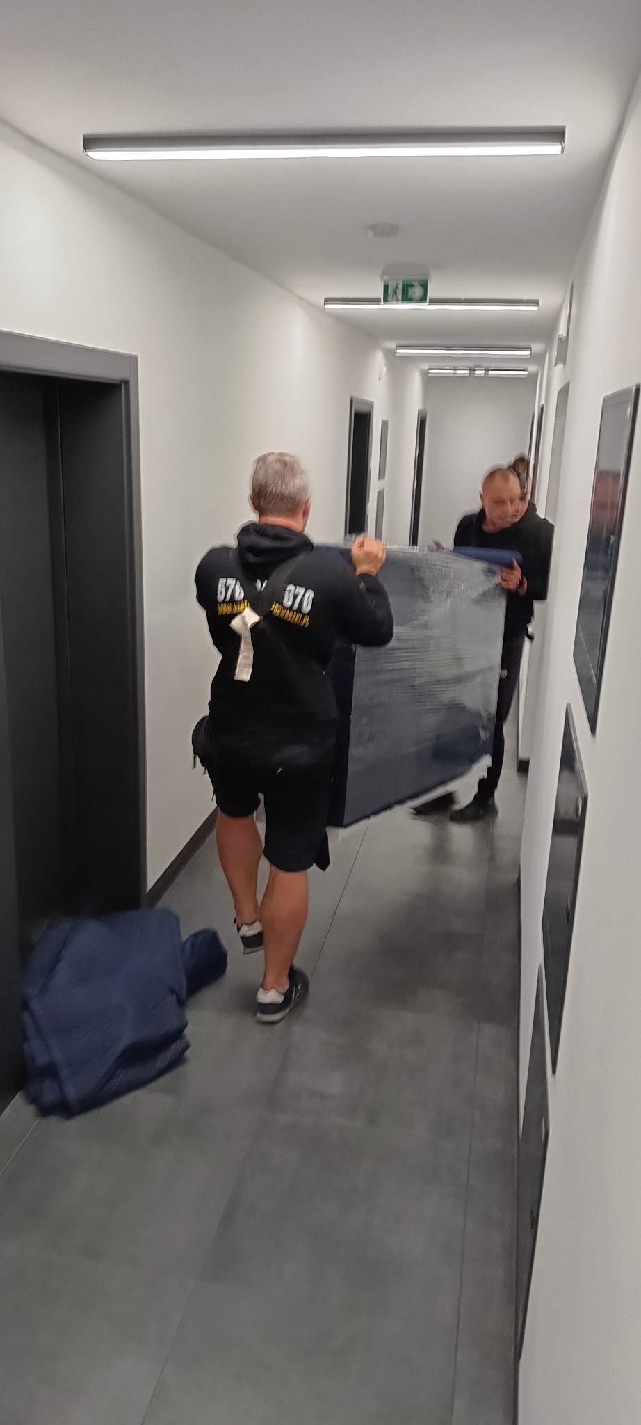 Dwóch mężczyzn niesie owinięty mebel w korytarzu, widoczne logo firmy na bluzie, co sugeruje profesjonalną przeprowadzkę w budynku.