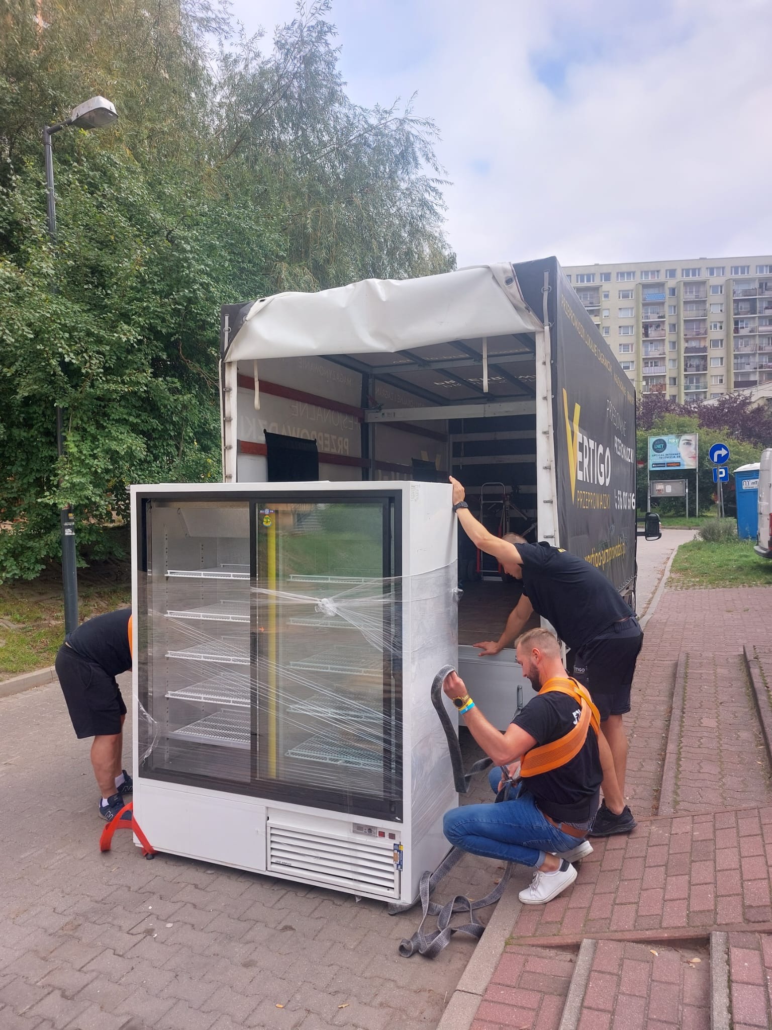 Dwóch mężczyzn w uprzężach transportowych ładuje owiniętą folią chłodziarkę do otwartej furgonetki firmy Vertigo na tle miejskiego krajobrazu.