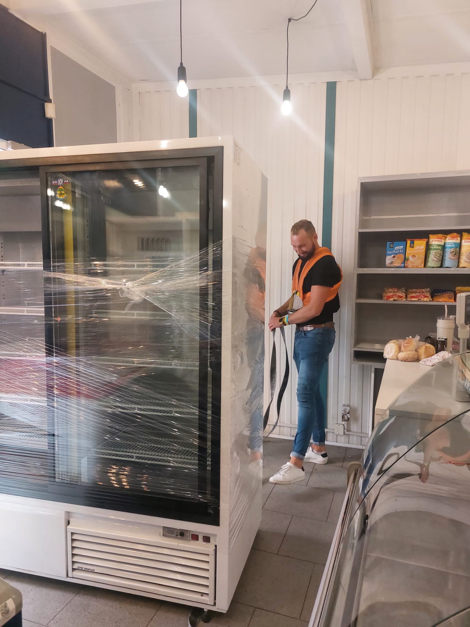 Pracownik w kamizelce zabezpieczającej mocuje pasy do owiniętej folią chłodziarki sklepowej przed transportem. W tle półki z produktami spożywczymi.