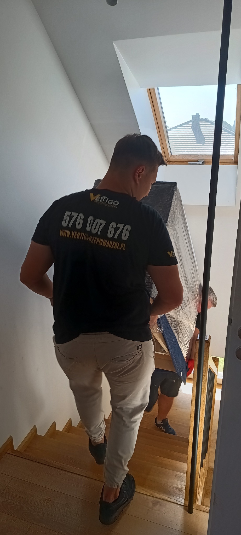Dwóch mężczyzn w koszulkach z logo firmy wnoszących zapakowany mebel po drewnianych schodach w jasnym wnętrzu z oknem dachowym.