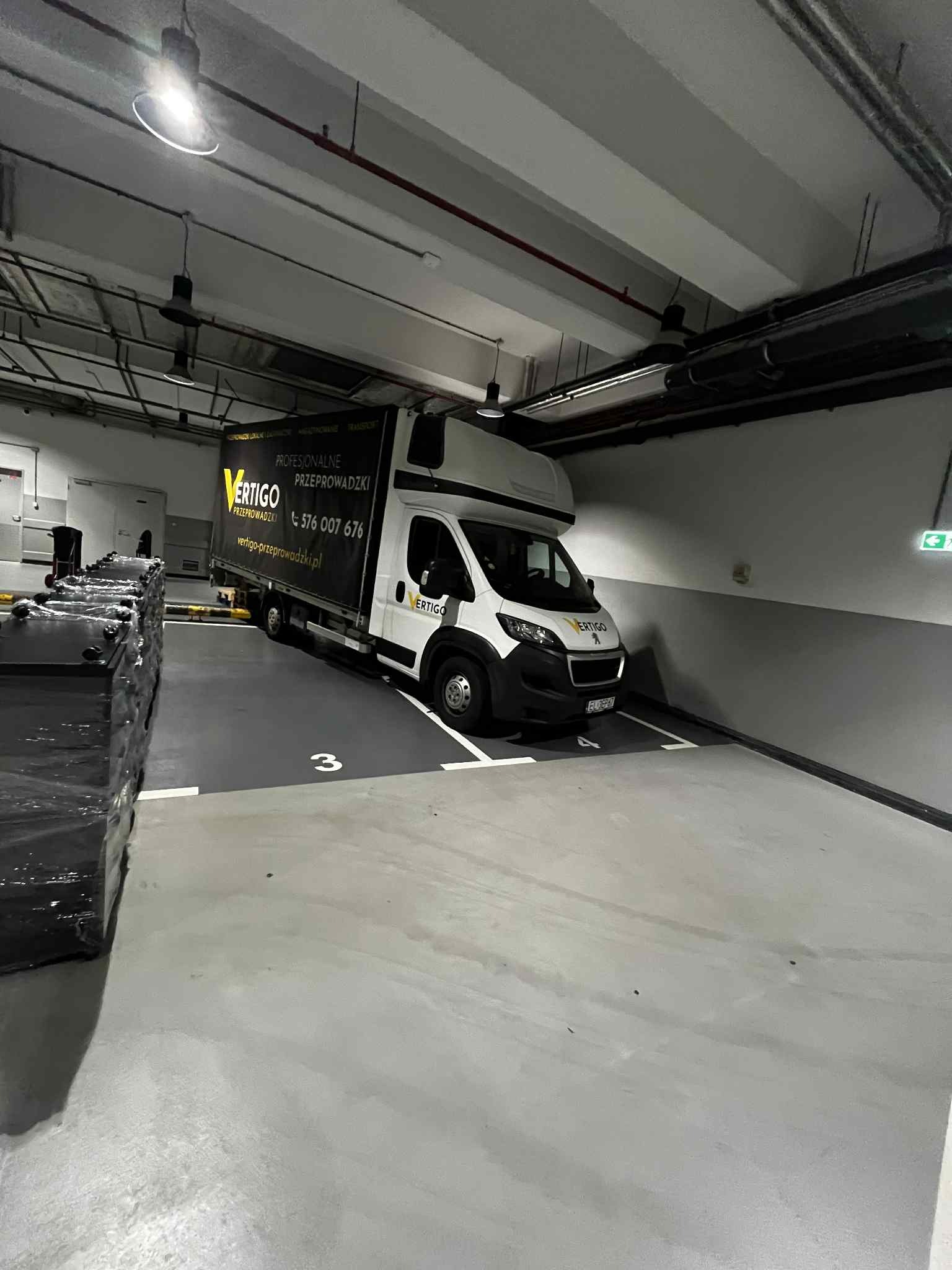 Biały bus Vertigo Przeprowadzki zaparkowany na numerowanym miejscu w garażu podziemnym, obok sterta spakowanych przedmiotów. Widoczne dane kontaktowe.