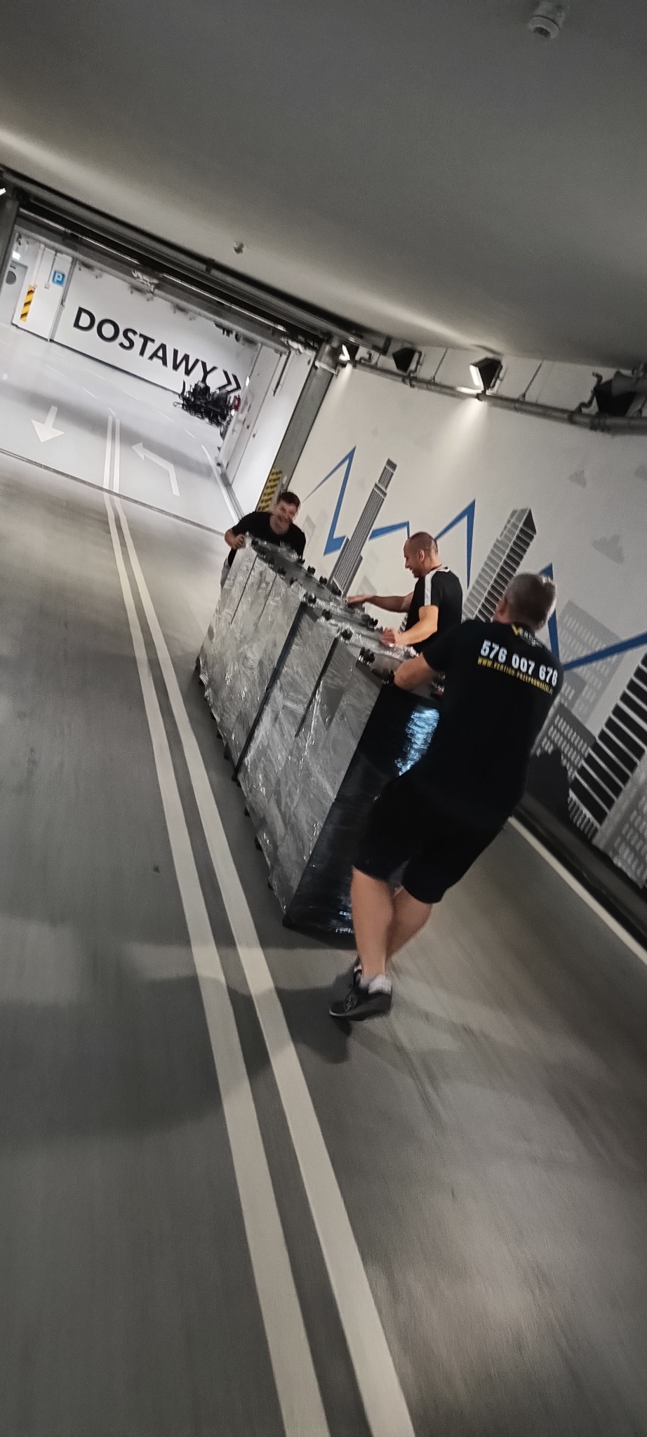 Trzech mężczyzn w czarnych koszulkach transportuje dużą, owiniętą folią paczkę w garażu z napisem 'Dostawy' i grafiką miasta.