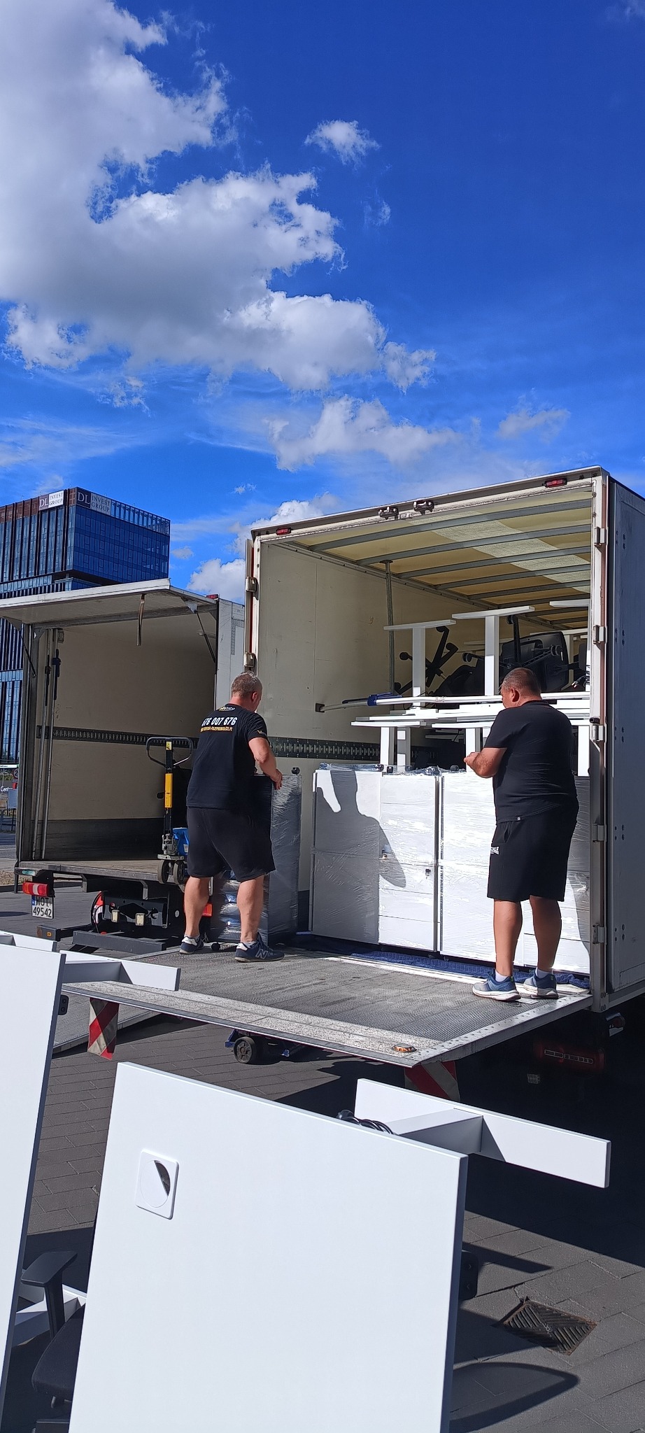 Dwóch mężczyzn ładuje białe, zapakowane meble biurowe do otwartej ciężarówki transportowej na tle błękitnego nieba z chmurami i nowoczesnego budynku.