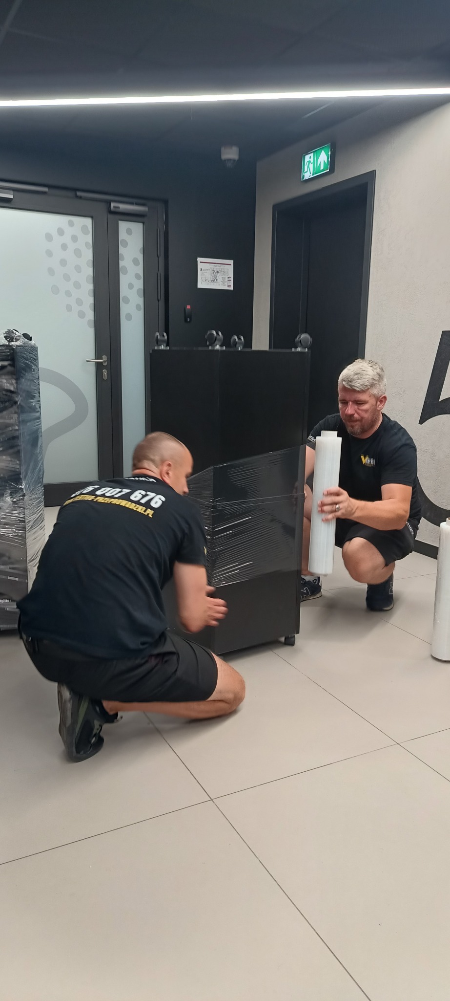 Dwóch mężczyzn w czarnych koszulkach owija czarny mebel folią stretch w jasnym pomieszczeniu. Przygotowanie do transportu, zabezpieczenie mienia podczas przeprowadzki.