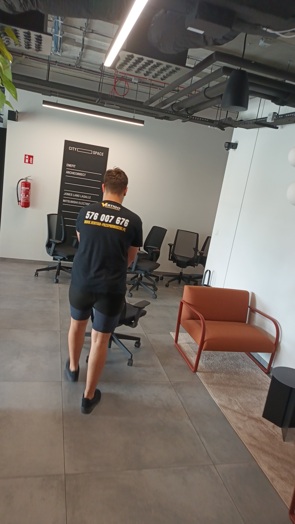 Pracownik w koszulce z logo firmy transportowej przesuwa krzesło biurowe w nowoczesnym biurze. Widoczne biurka, krzesła i elementy wystroju wnętrz.