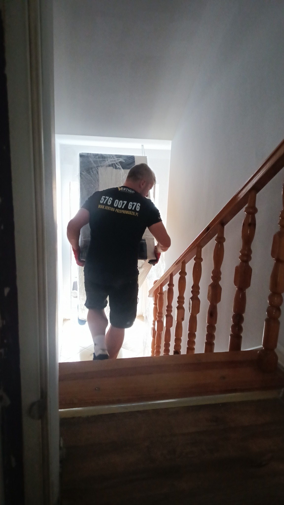 Pracownik firmy przeprowadzkowej w czarnej koszulce z logo, wnoszący zapakowany mebel po schodach w jasnym wnętrzu domu. Widoczne drewniane schody i fragment korytarza.