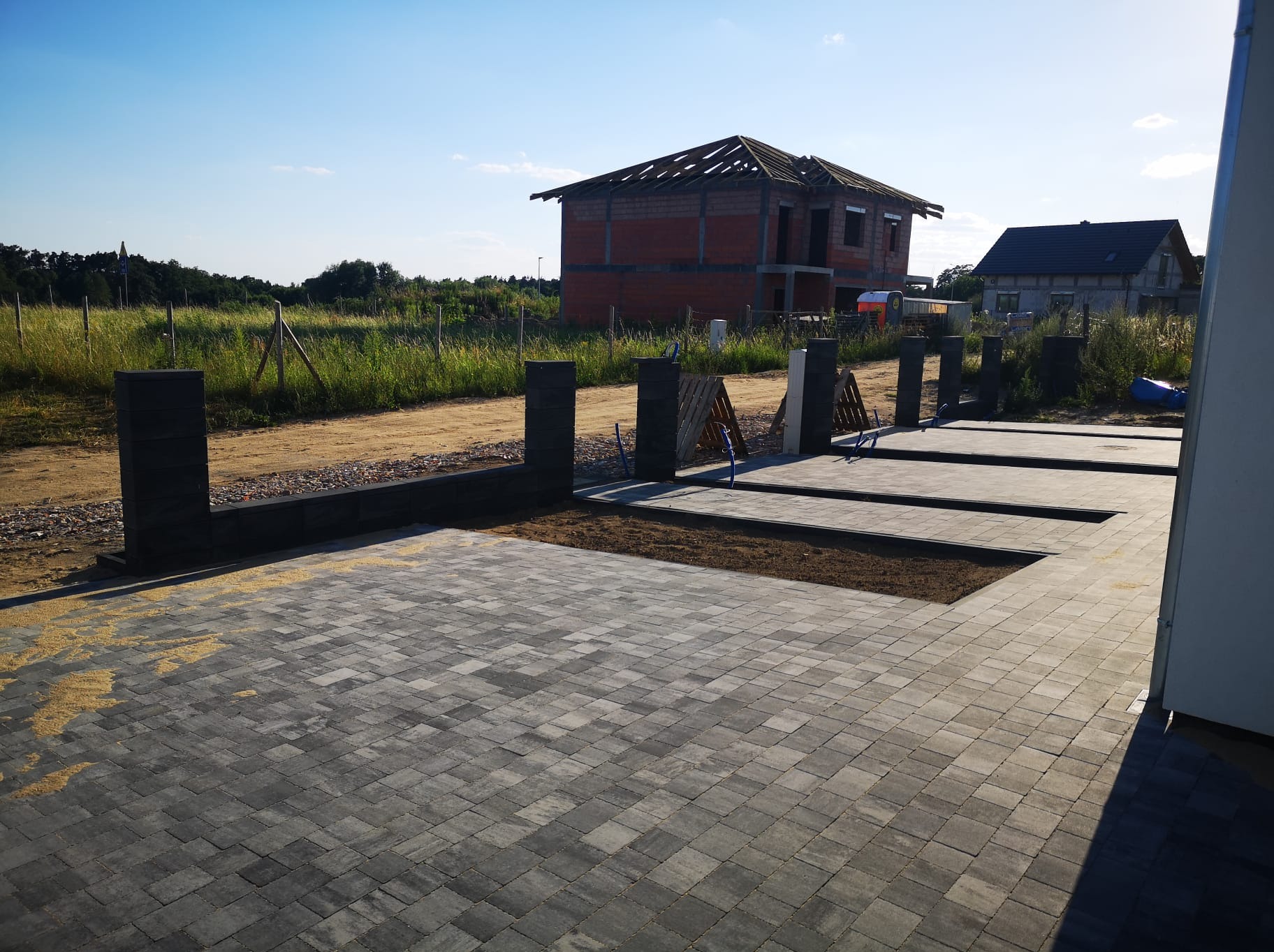 Ułożona szara kostka brukowa na podjeździe, widoczne słupki ogrodzeniowe i w tle budynek w trakcie budowy.