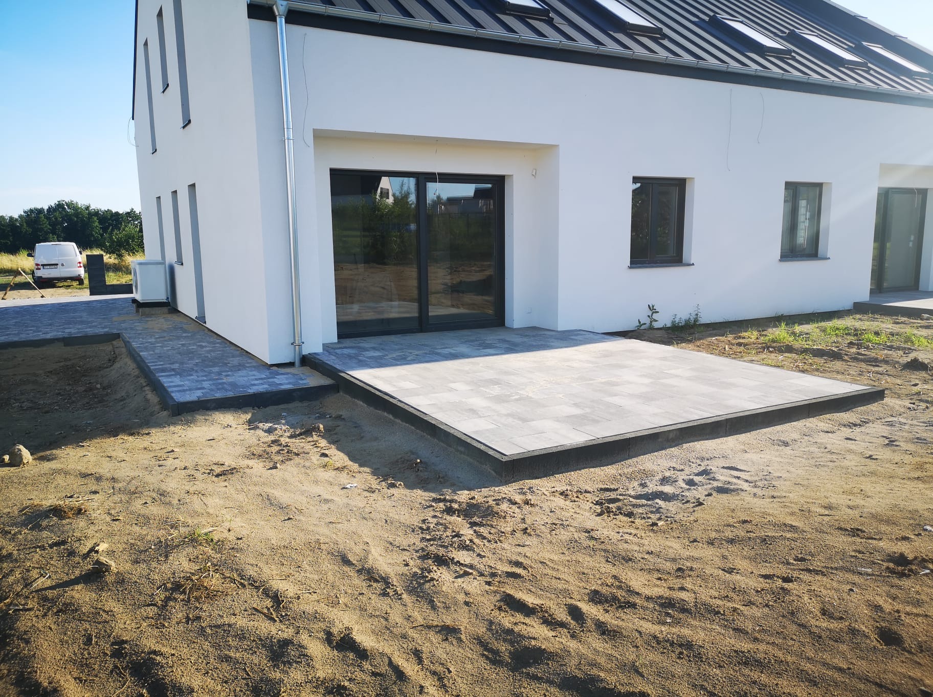 Nowoczesny taras z szarej kostki brukowej przed białym domem, widoczny fragment chodnika z kostki i przygotowany teren pod trawnik.