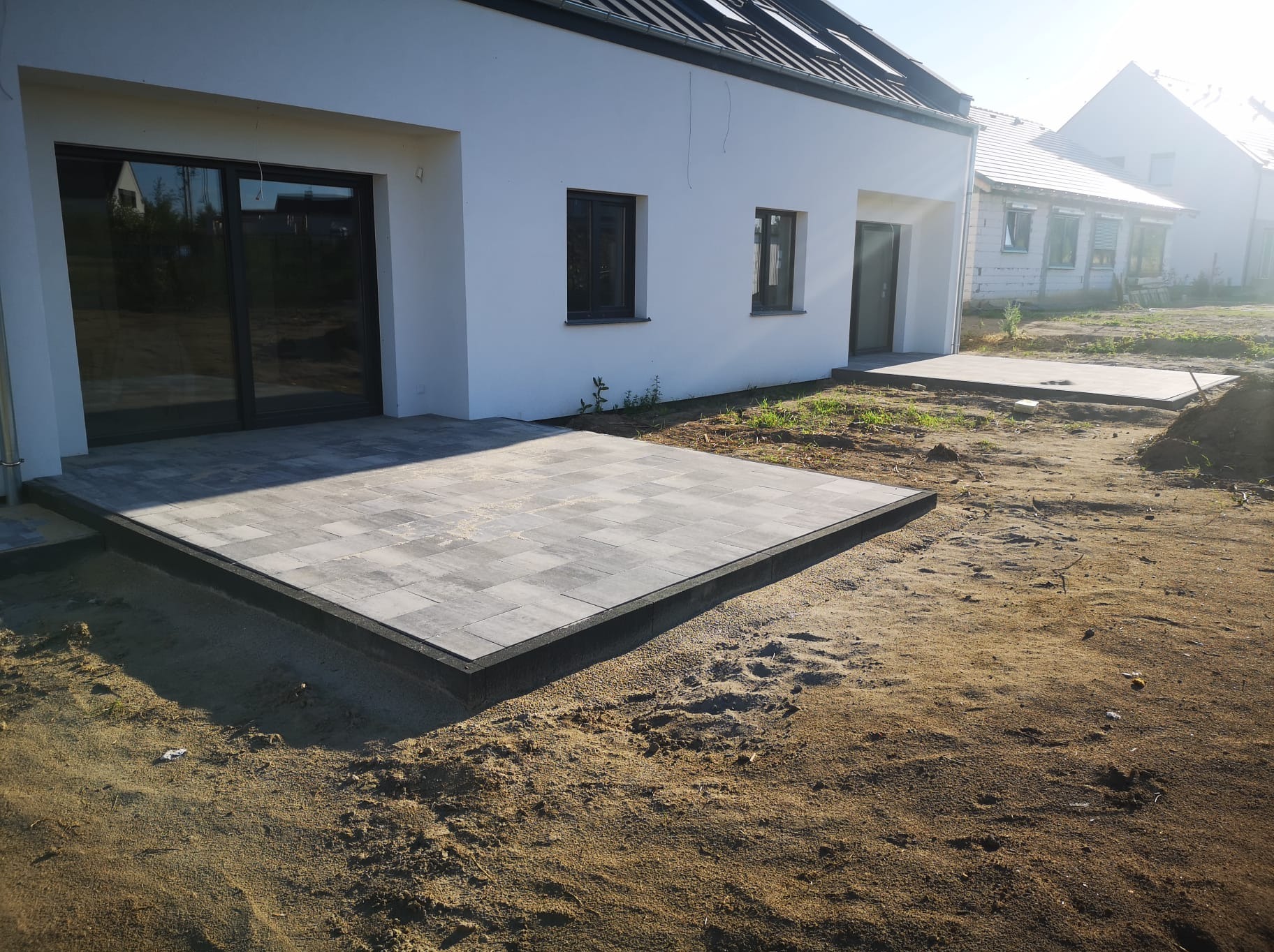 Nowoczesny taras z szarej kostki brukowej ułożony przy białym domu jednorodzinnym, widoczne obrzeża i przygotowane podłoże z piasku.