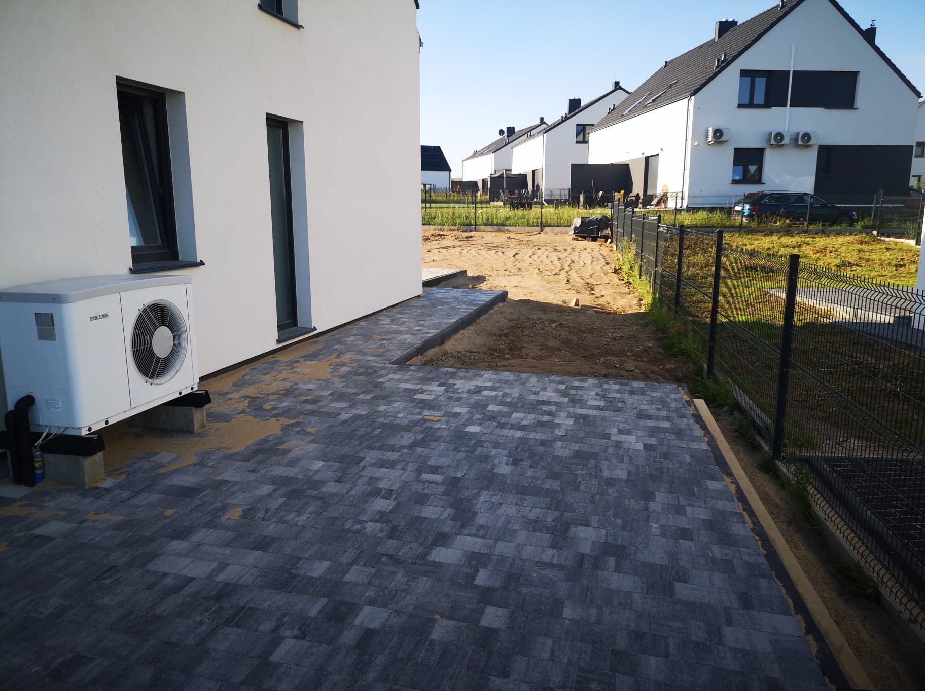 Ułożona szara kostka brukowa na podjeździe nowego domu z białymi ścianami, widoczny agregat klimatyzacji i teren przygotowany pod dalsze prace brukarskie.