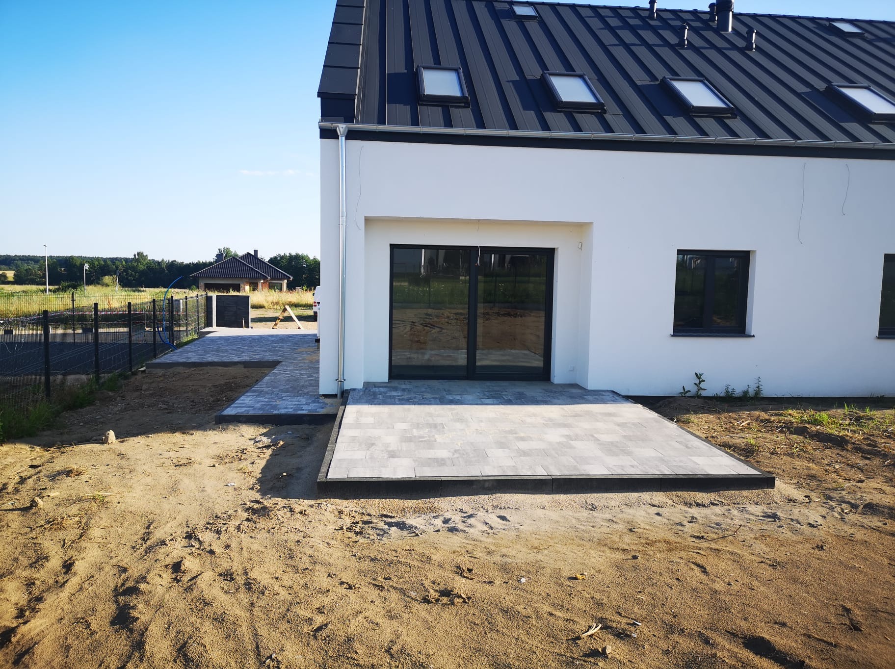 Nowoczesny dom z białymi ścianami i antracytowym dachem, z wybrukowanym szarym brukiem tarasem i chodnikiem, widok na trawnik i ogrodzenie w tle.