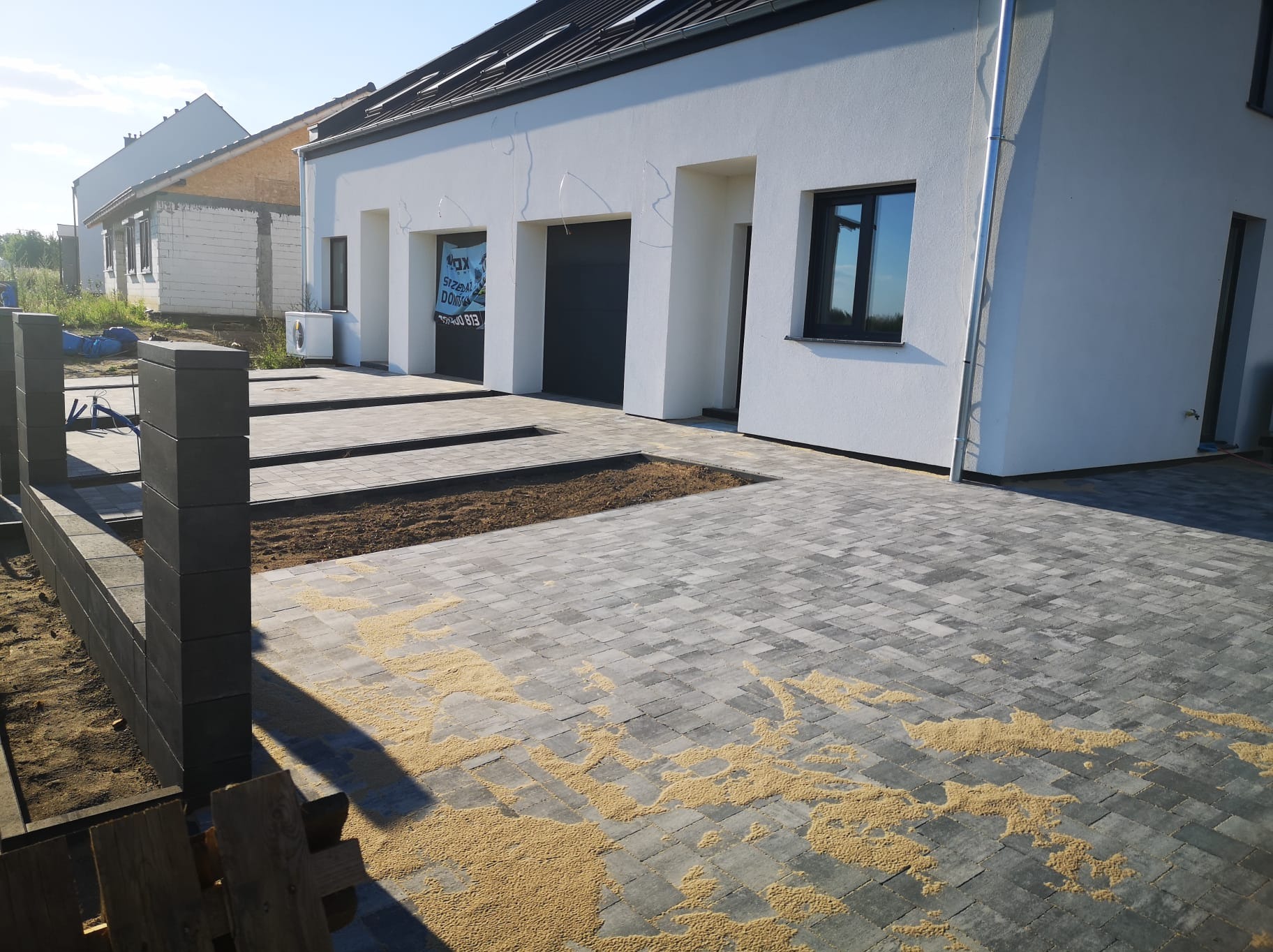 Ułożona szara kostka brukowa przed nowym białym domem z czarnymi oknami i bramami garażowymi, z widocznymi pozostałościami piasku spoinującego na powierzchni i przygotowanymi rabatami na rośliny.