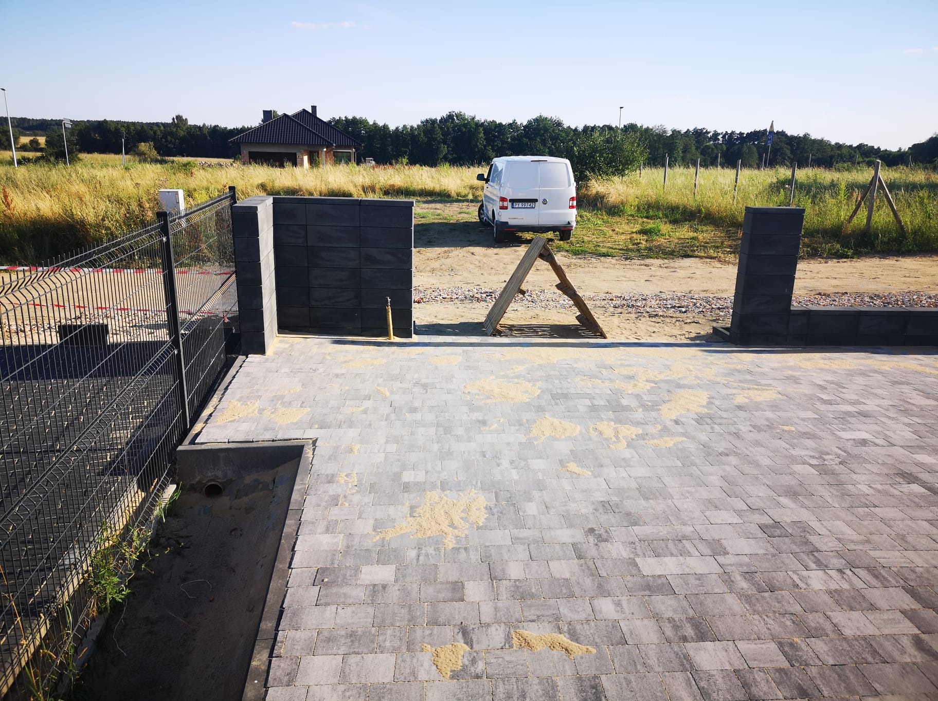 Ułożona szara kostka brukowa z piaskiem na powierzchni, widoczny fragment ogrodzenia panelowego, słupki z betonowych bloczków, biały bus w tle i drewniany kozioł.