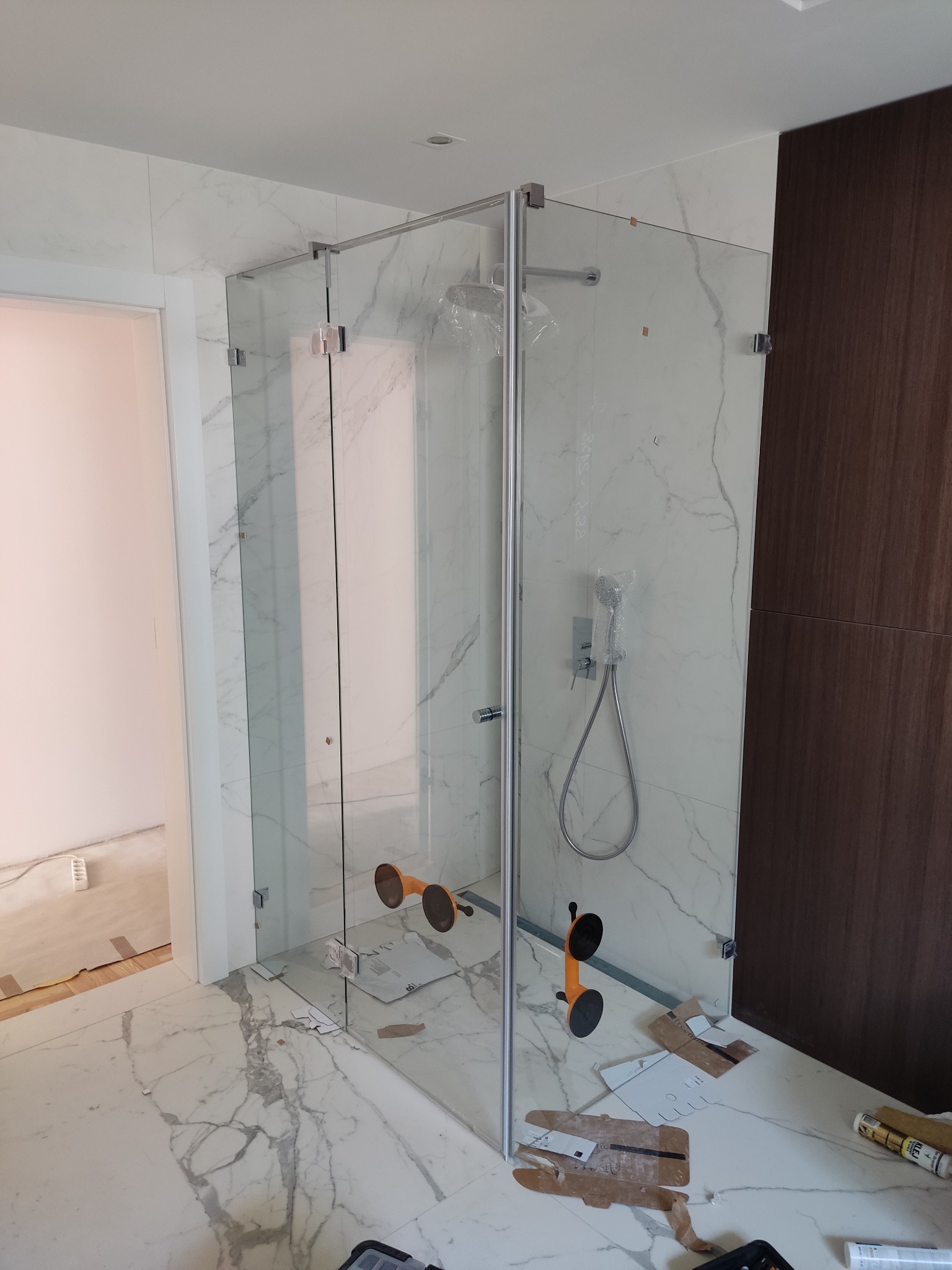 Montaż kabiny prysznicowej ze szkła hartowanego, widoczne przyssawki montażowe, marmurowe wykończenie ścian i podłogi, oraz częściowo zamontowana armatura.