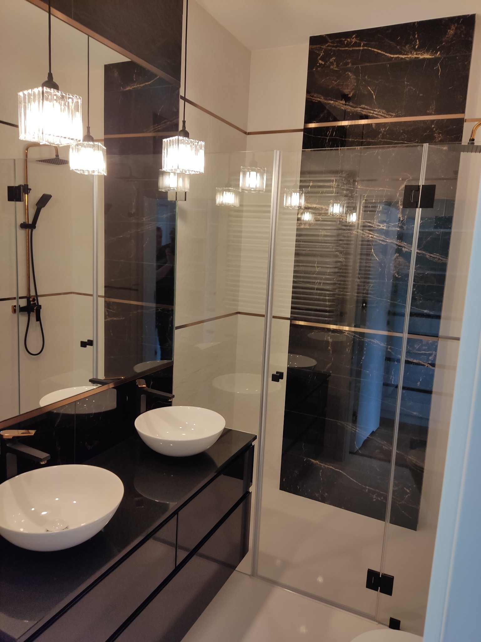 Nowoczesna łazienka z dwoma białymi umywalkami na czarnym blacie, przeszklona kabina prysznicowa z czarnymi elementami i marmurową ścianą, oświetlenie z kryształowymi kloszami.
