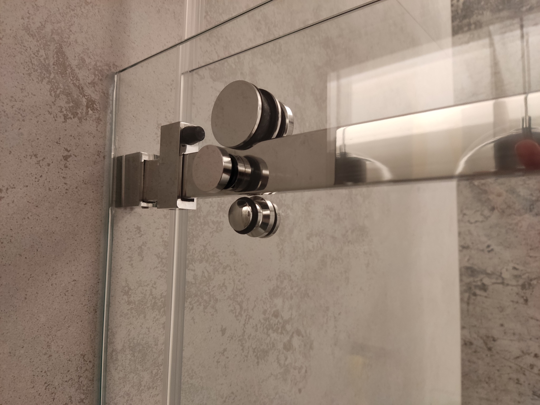 Szklana kabina prysznicowa z przesuwnymi drzwiami, widoczne metalowe okucia i rolki na tle ściany w odcieniach szarości.