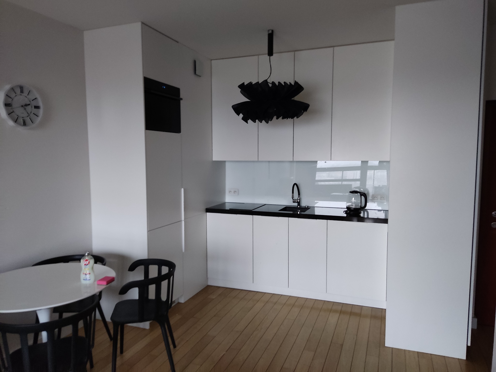 Minimalistyczna kuchnia z białymi szafkami, czarnym blatem, drewnianą podłogą i czarnym designerskim żyrandolem, widoczny stół z krzesłami i płynem do naczyń.