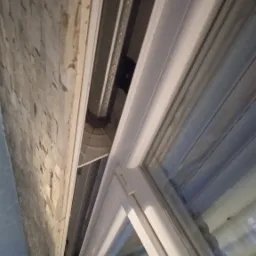 Mechanizm rolety zewnętrznej zamontowany nad oknem z białymi ramami, widoczny od spodu, z fragmentem kamiennej ściany po lewej stronie.