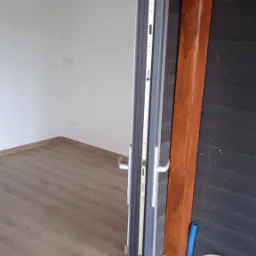 Otwarta aluminiowa rama okienna z widokiem na wnętrze pomieszczenia z drewnianą podłogą, na zewnątrz widoczna drewniana konstrukcja i kamienny chodnik, obok ramy stoi butelka środka czyszczącego.