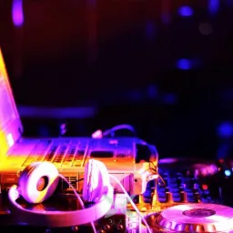 Laptop, słuchawki i mikser DJ podświetlone fioletowym i pomarańczowym światłem scenicznym.