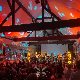 Orkiestra smyczkowa gra koncert dla publiczności w sali z drewnianą konstrukcją dachu i czerwono-niebieskim oświetleniem, obrazy wiszą na ścianach.