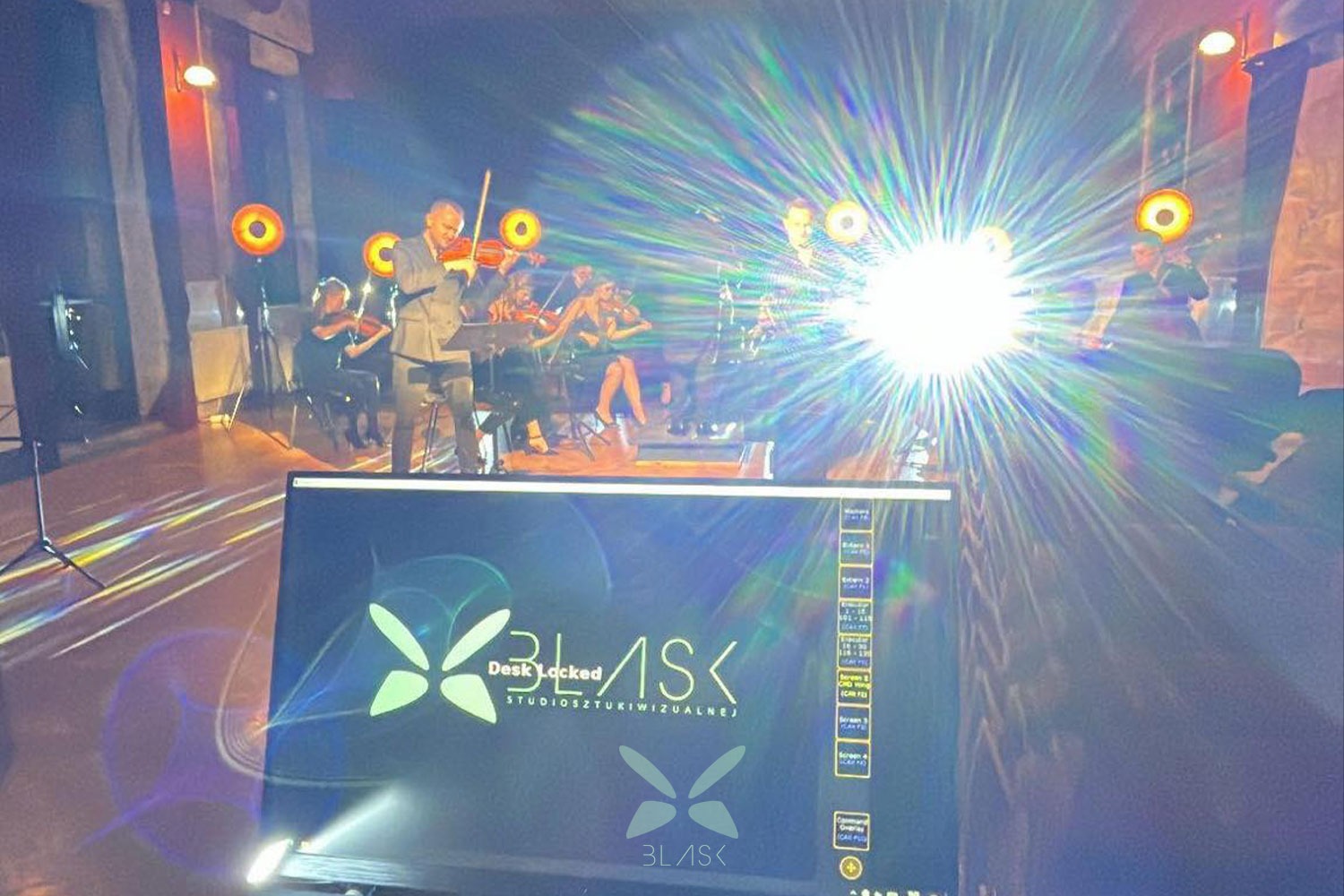 Zespół muzyczny z skrzypcami na scenie, oświetlony reflektorami, widok zza ekranu komputera z logiem '3LASK Studio Sztuki Wizualnej'.
