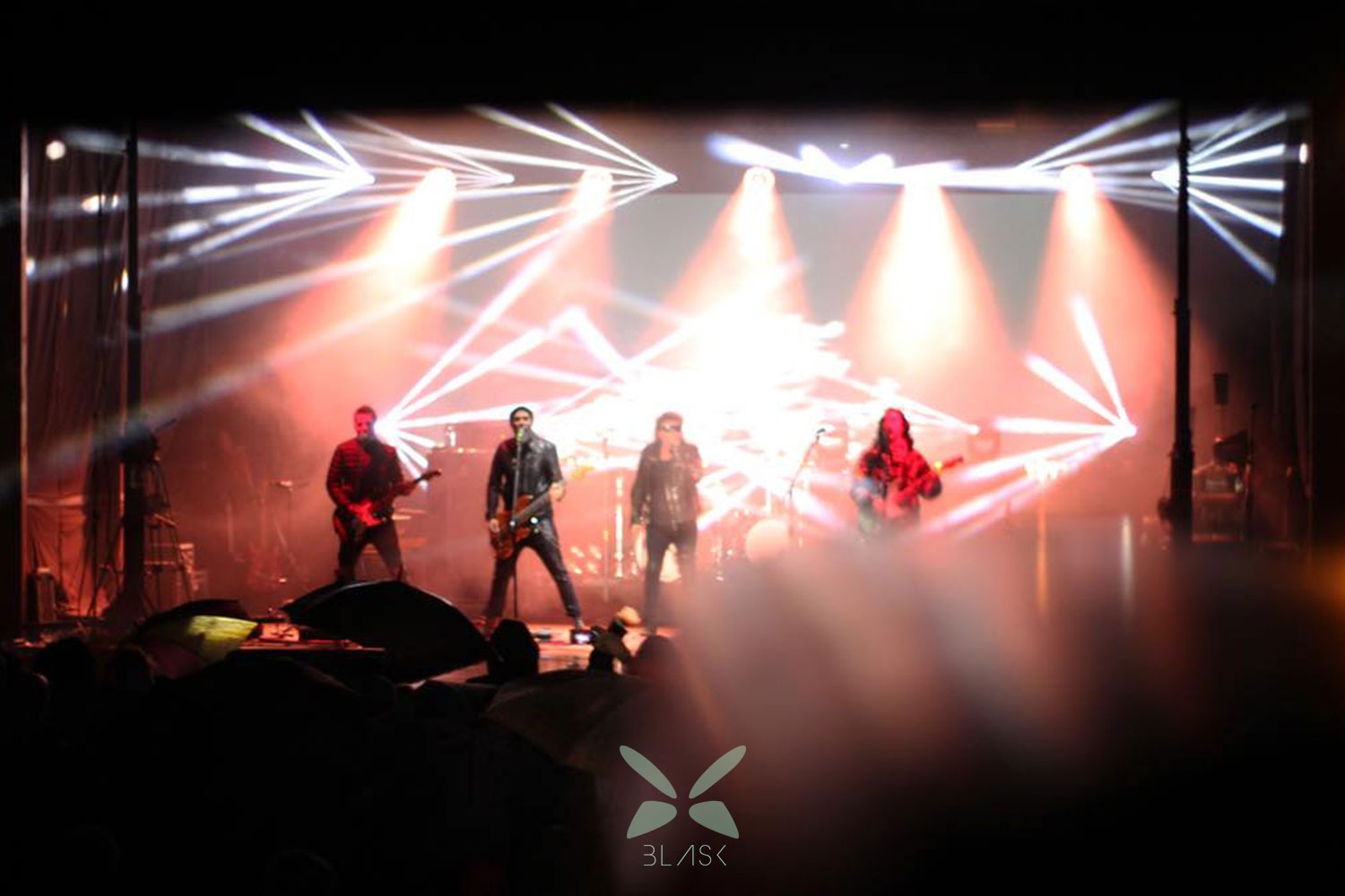 Zespół muzyczny na scenie w trakcie występu, dynamiczne oświetlenie sceniczne, widoczny tłum na widowni