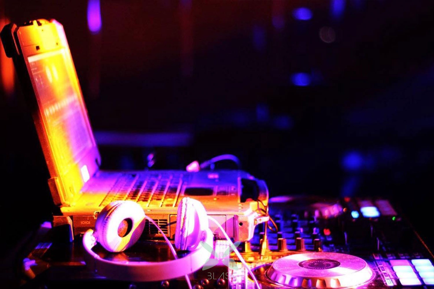 Laptop, słuchawki i mikser DJ podświetlone fioletowym i pomarańczowym światłem scenicznym.