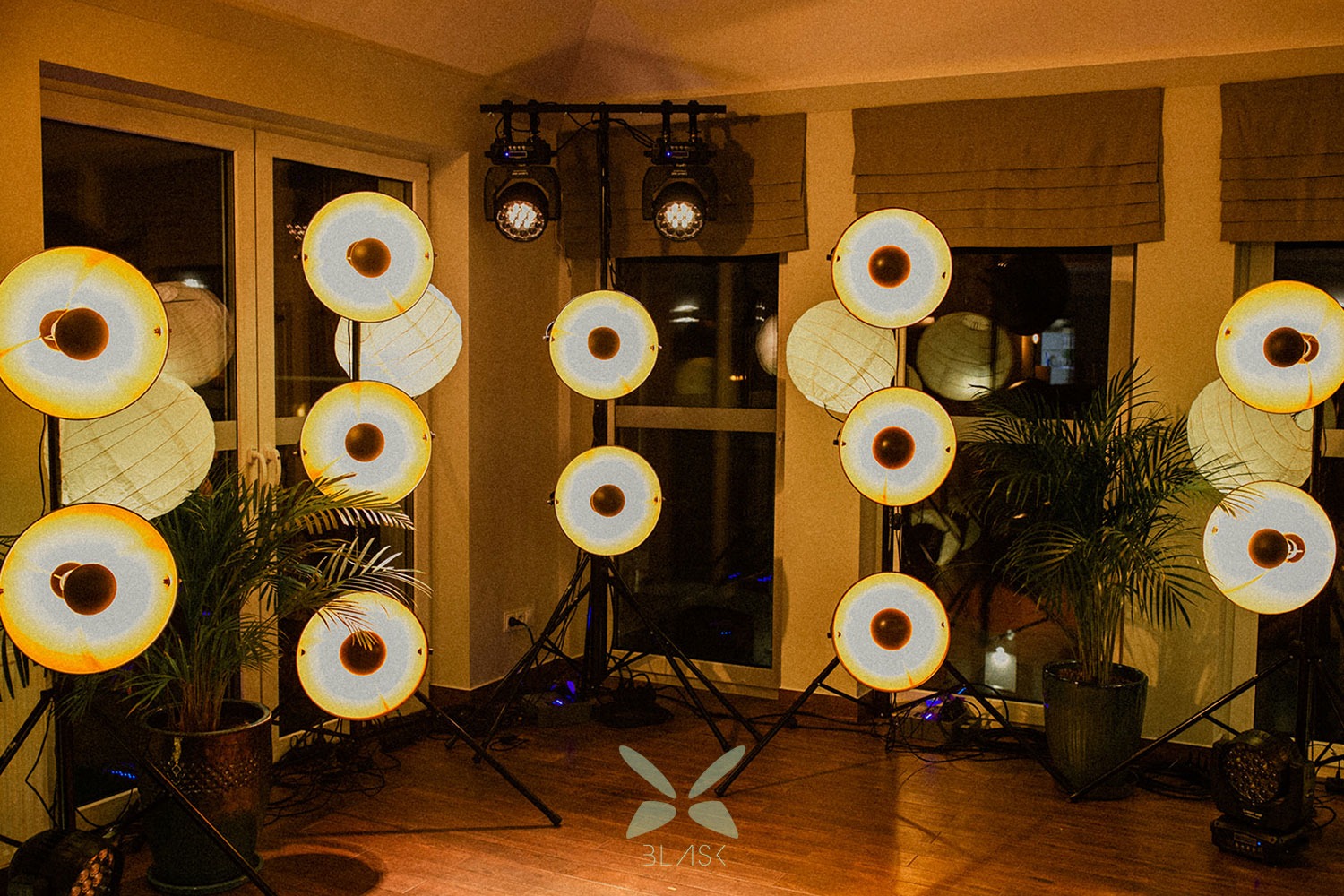 Okrągłe lampy dekoracyjne na czarnych statywach, oświetlające wnętrze z parkietem i roślinami doniczkowymi, dodatkowe reflektory na górze, widoczne logo na podłodze.