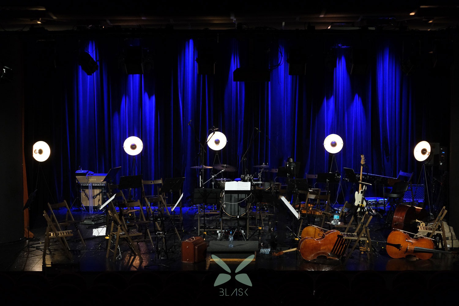 Scena przed koncertem: instrumenty smyczkowe, gitara, perkusja, keyboard, krzesła i lampy oświetleniowe na tle niebieskich zasłon.