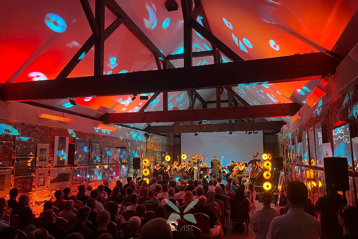 Orkiestra smyczkowa gra koncert dla publiczności w sali z drewnianą konstrukcją dachu i czerwono-niebieskim oświetleniem, obrazy wiszą na ścianach.