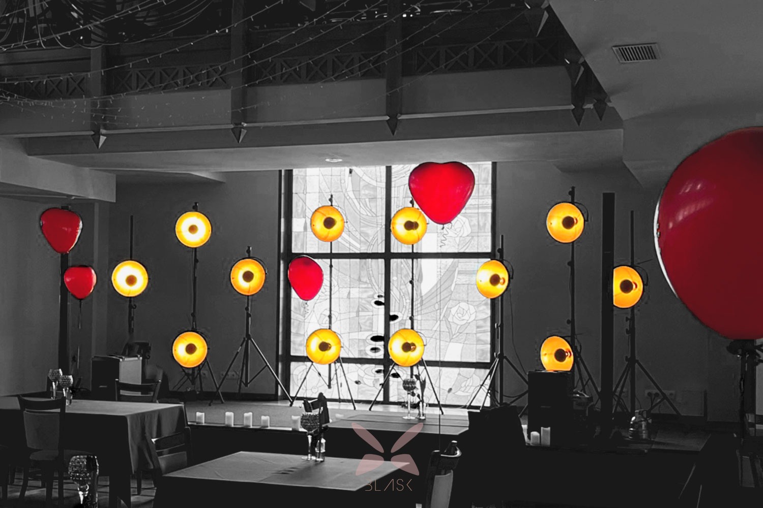 Ustawienie sceniczne w Nowym Sączu z reflektorami na statywach i czerwonymi balonami w kształcie serc, na tle okna z witrażem, w sali z nakrytymi stołami.