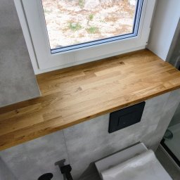 Make Me Woody Konrad Klimiuk - Blat z jasnego drewna pod oknem w łazience, widok na zewnątrz, minimalistyczna aranżacja z szarą płytką i czarnym przyciskiem spłuczki.