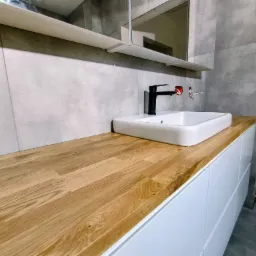 Łazienka w trakcie wykańczania: drewniany blat z umywalką nablatową i czarną armaturą, szare płytki na ścianie, widoczne kable elektryczne.