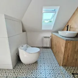 Aranżacja łazienki na poddaszu ze skośnym oknem dachowym, białą ceramiką i szafką z drewnianym blatem. Geometryczne płytki podłogowe.