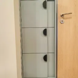 Szara szafka na buty z trzema poziomami i półokrągłymi uchwytami, zamontowana obok drewnianych drzwi wejściowych. Minimalistyczny design, jasne wnętrze.