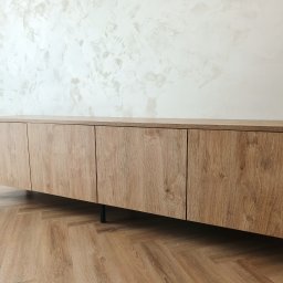 Make Me Woody Konrad Klimiuk - Nowoczesna szafka RTV z drewnianymi frontami i czarnymi nóżkami, na tle ściany z dekoracyjnym tynkiem. Ujęcie z niskiej perspektywy, podkreślające fakturę drewna.