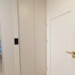 Make Me Woody Konrad Klimiuk - Jasna szafa wnękowa z minimalistycznymi, czarnymi uchwytami, wkomponowana w korytarz z parkietem ułożonym w jodełkę. Obok białe drzwi z złotą klamką.