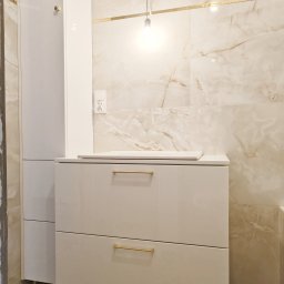 Make Me Woody Konrad Klimiuk - Biała szafka łazienkowa z dwiema szufladami i złotymi uchwytami, na tle marmurowych płytek i wiszącej żarówki. Element zabudowy łazienki.