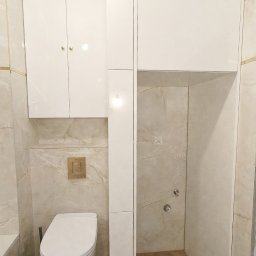 Make Me Woody Konrad Klimiuk - Biała zabudowa łazienkowa z szafkami nad toaletą i wnęką prysznicową. Beżowe płytki imitujące marmur, drewnopodobna podłoga ułożona w jodełkę.