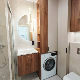 Make Me Woody Konrad Klimiuk - Nowoczesna łazienka z drewnianą szafką pod umywalką, okrągłym lustrem z podświetleniem LED i pralką Electrolux w zabudowie. Marmurowe płytki na ścianach i podłodze.
