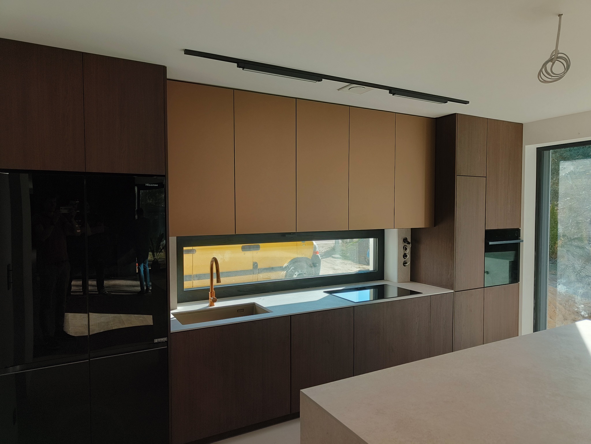 Nowoczesna kuchnia z brązowymi i beżowymi frontami, minimalistycznym oświetleniem szynowym i miedzianą baterią, widok z okna na żółty samochód