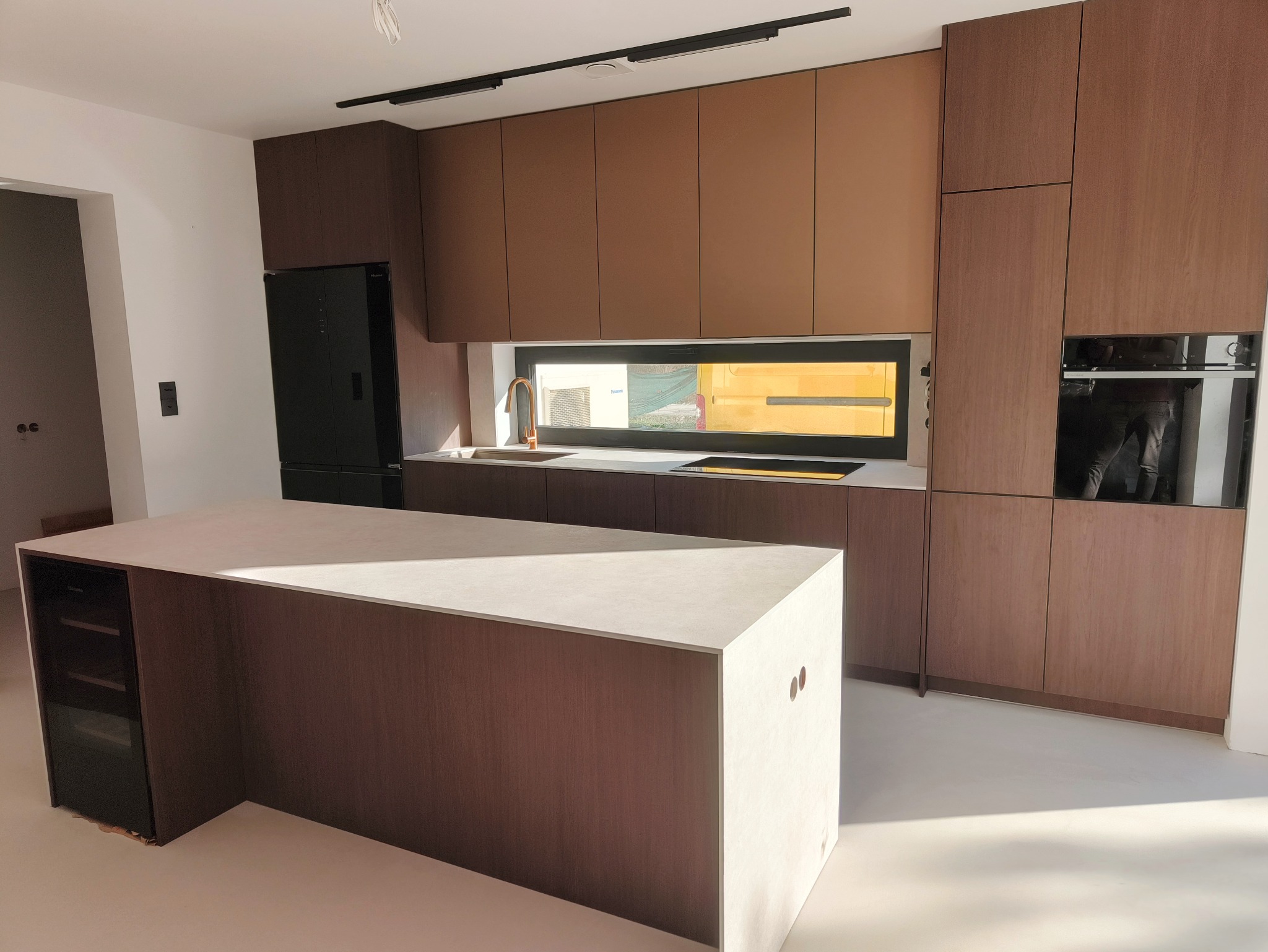 Nowoczesna kuchnia z brązowymi szafkami, wyspą z blatem w kolorze jasnego kamienia i czarną lodówką, oświetlona światłem słonecznym.