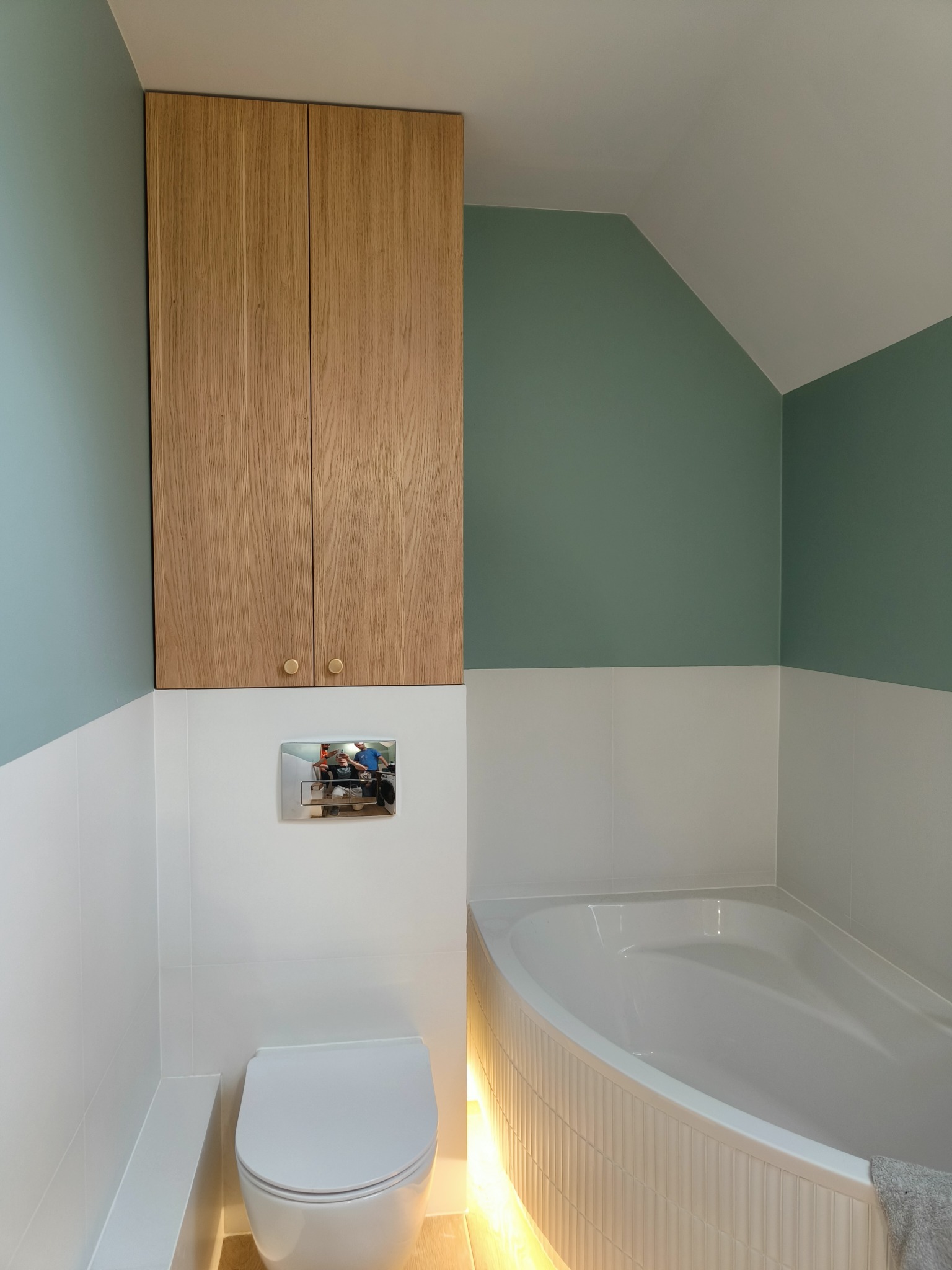 Nowoczesna łazienka z drewnianą szafką nad toaletą, białą armaturą i wanną z podświetleniem LED. Ściany wykończone w bieli i odcieniach zieleni.