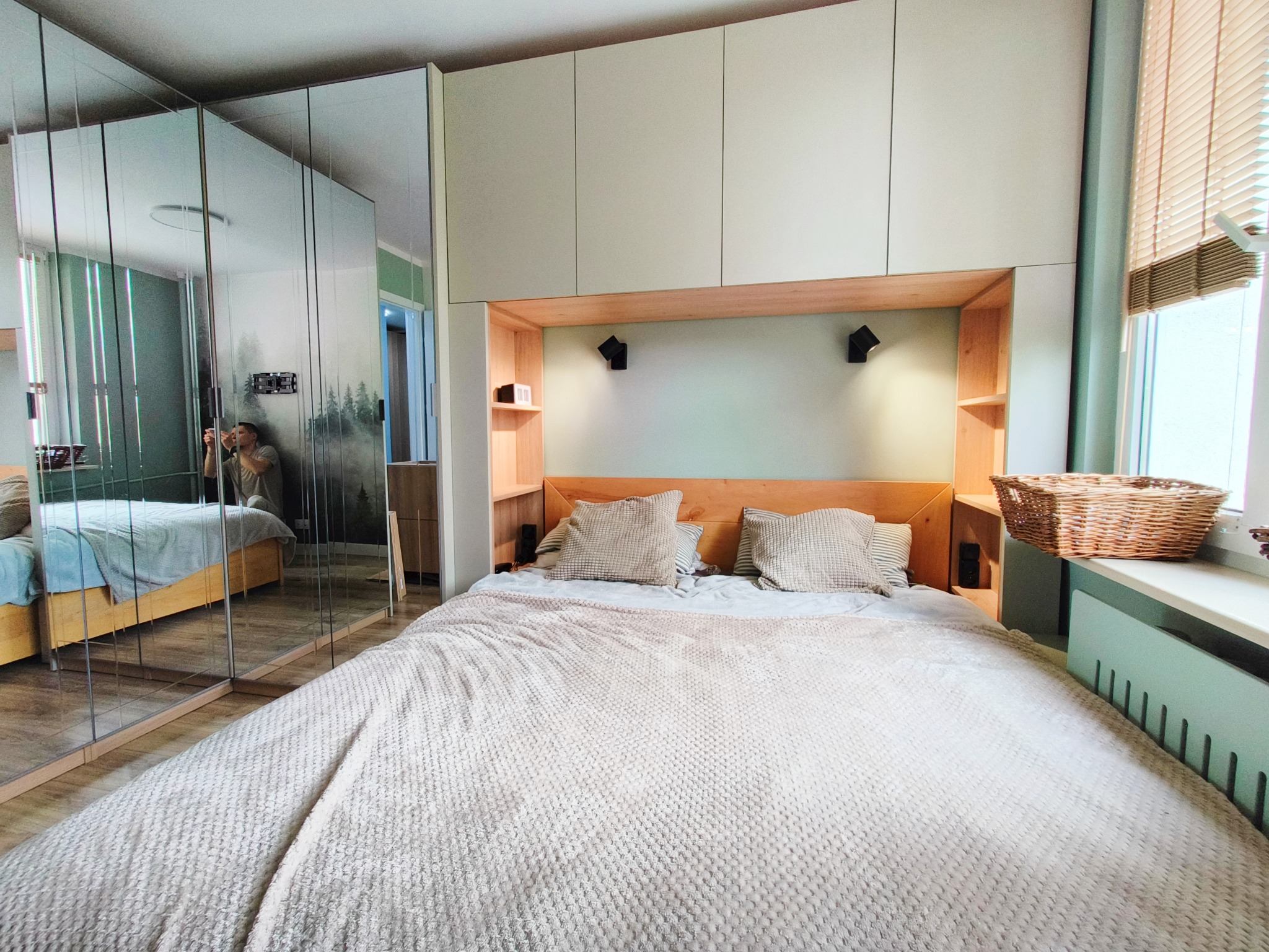 Nowoczesna sypialnia z łóżkiem, zabudową nad nim i szafą z lustrami. Widoczny kosz wiklinowy i osoba robiąca zdjęcie w odbiciu lustra.